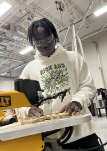 A student in a white sweatshirt uses a bandsaw on his wood project.