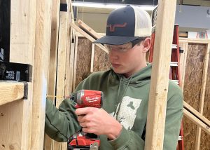 A student in a baseball cap and safety goggles uses a power drill while in the Building Trades program.