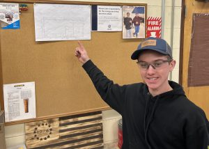 A student in a baseball cap and safety points up at a set of blueprints while in the Building Trades program.