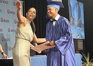 A woman gestures towards the sky after greeting a graduate on stage at a graduation ceremony.