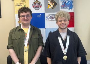 Two students are smiling at the camera, standing next to each other and each one is wearing a medal around the neck.