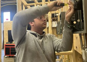 A student wearing safety glasses is standing in front of an electrical box on the wall using a screwdriver to install a component.