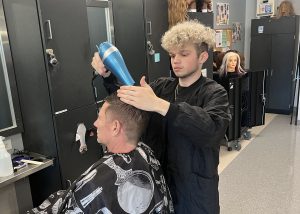 A student is standing over an adult sitting in a chair blow drying the adult's hair after giving a hair cut.
