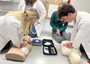 Four students are kneeling on the floor practicing how to administer CPR to practice dummies which are lying on the floor.