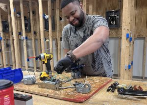 A student wearing gloves is standing behind a work space working on an electrical wiring harness.