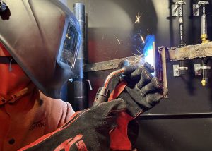 A student iwearing protective head gear, clothing, and gloves, is welding two pieces of metal together.