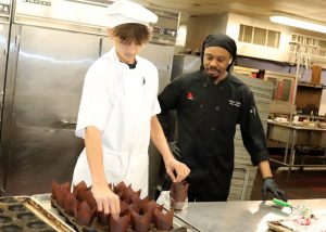 A student dressed in chef clothing is placing liners in a cupcake tin.