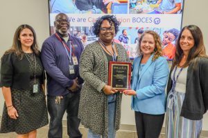 Five people stand in a row. The person in the center is holding a plaque. They are all looking at the camera smiling. Behind them is a screen with several images, with text that reads We are Capital Region BOCES