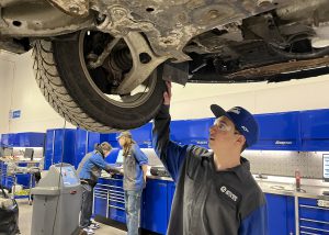 A student is standing under a car that is on a lift inspecting how a tire is attached with their hand touching the tire's tread.