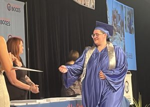 A student wearing a gradutation cap and gown reaches out with their hand to receive a document.