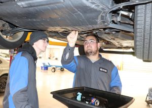 Two students are standing under an automobile that is on a lift looking at the underbody.