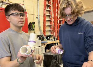 One student holds up PVC pipe while another applies glue to his PVC pipe