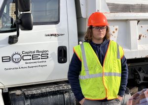 Student with long hair, orange helmet and reflective vest stands in front of a BOCES dump truck