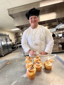 A student smiles behind cupcakes he made