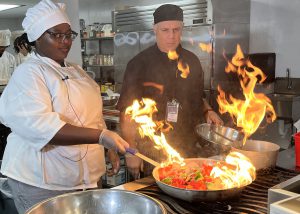 Culinary student and teacher cooking vegetables with flames coming from the pan