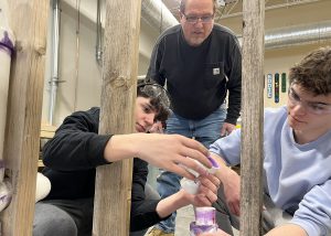 Two Plumbing Technology students work gluing PVC pipe pieces together between 2x4 studs while an instructor oversees their work.