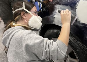 Female student in mask sanding Bondo on a car