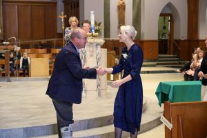 The setting is a church, two people are shaking hands. The people in the background are applauding