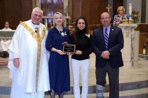 Four people are standing side by side. They are in a church. They are smiling at the camera. One is a priest, wearing a vestment that is white with gold trim. The two in the middle are holding a plaque; all are smiling