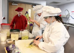 A prospective culinary student in red watches two white-smocked current students prepare vegetables