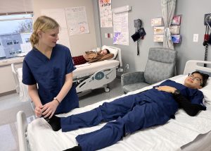 A nursing student smiles while manipulating the foot of another student who is lying in bed